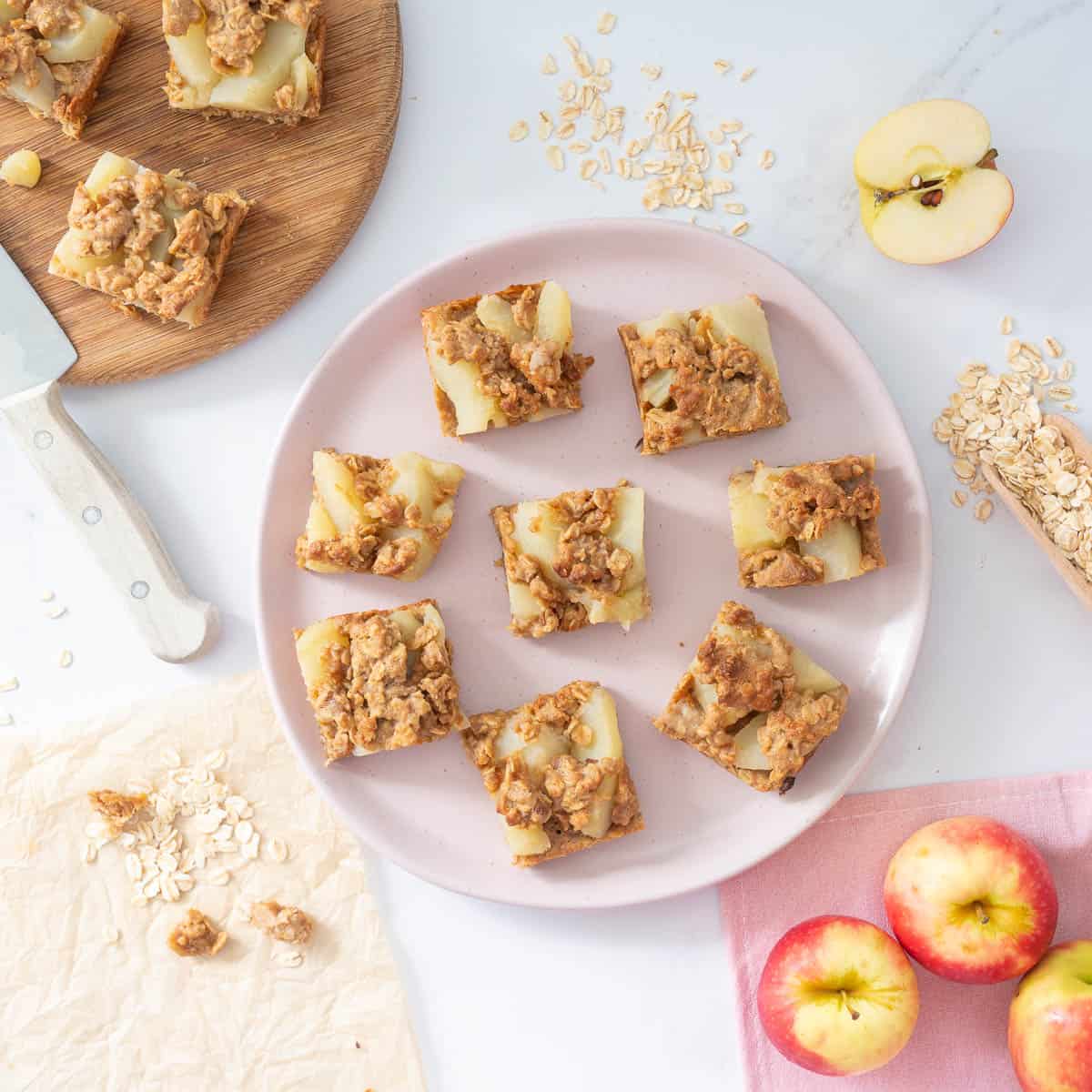 Eight pieces of apple slice on a pink plate surrounded by rolled oats and fresh apples on a bench top.