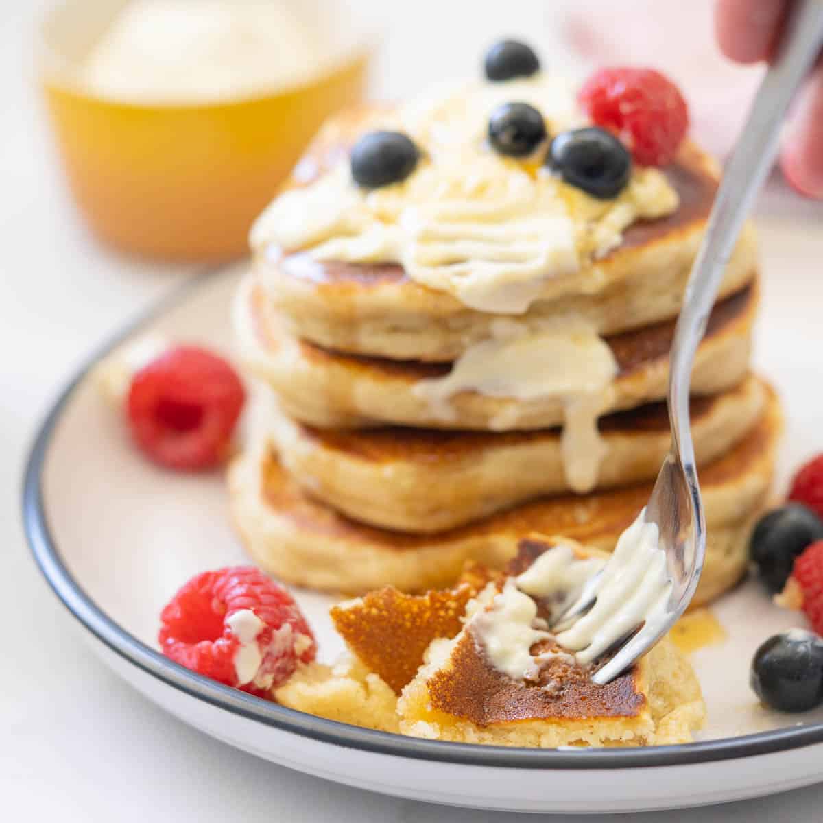 A fork taking a forkfull of pancakes, berries and yoghurt in front of a stack of pancakes.