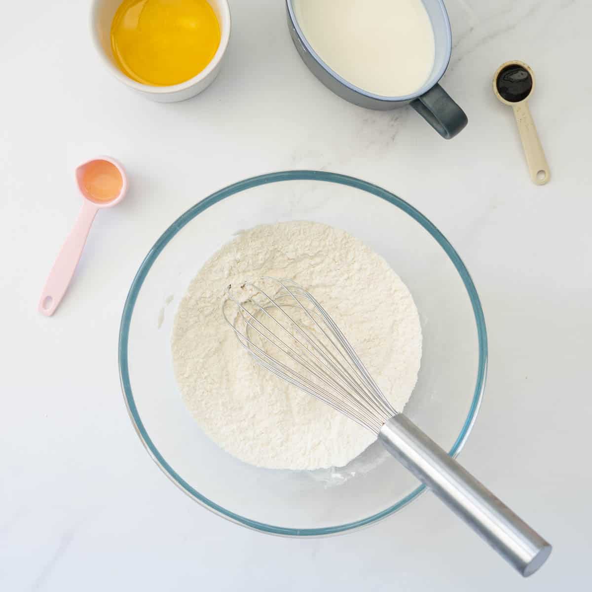 A large glass mixing bowl of dry ingredients with a whisk. 