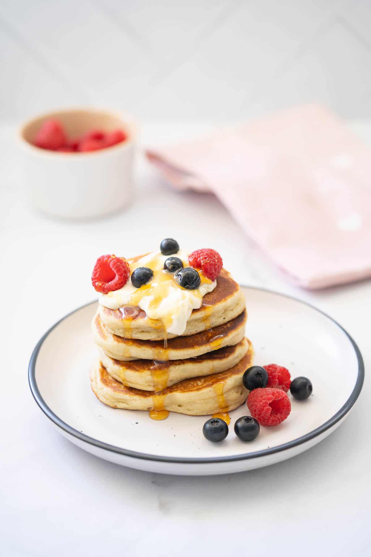Four thick pancakes stacked on a tower topped with yoghurt, berries and yoghurt with a bowl of raspberries and a pink tea towel in the background.