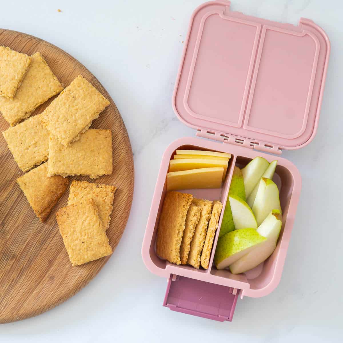 A pink bento snack box packed with crackers, cheese and slices of pear. 