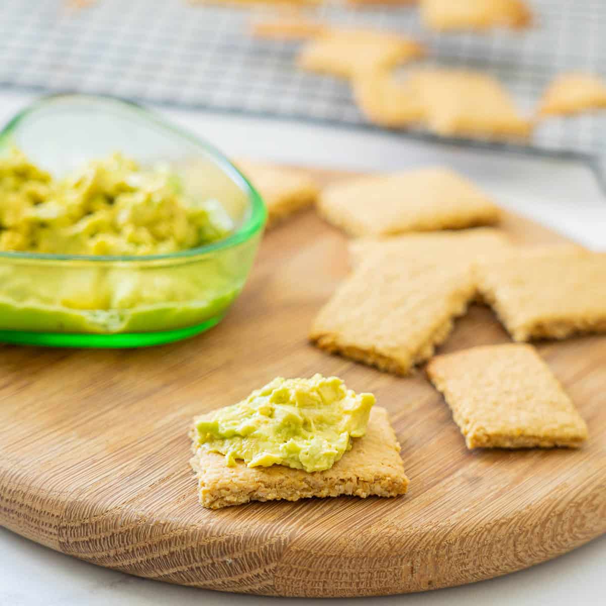 A cracker topped with avocado on a wooden board surrounded by other crackers. 