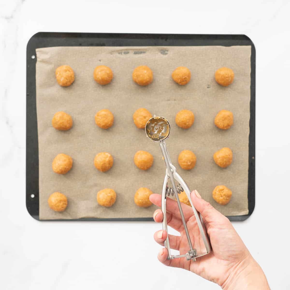A cookie scoop held above a baking tray of cookie dough balls ready to be pressed. 