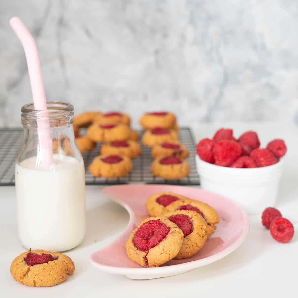 Three raspberry thumbprint cookies on a pink crescent shaped plate with a small bottle of milk to the side.