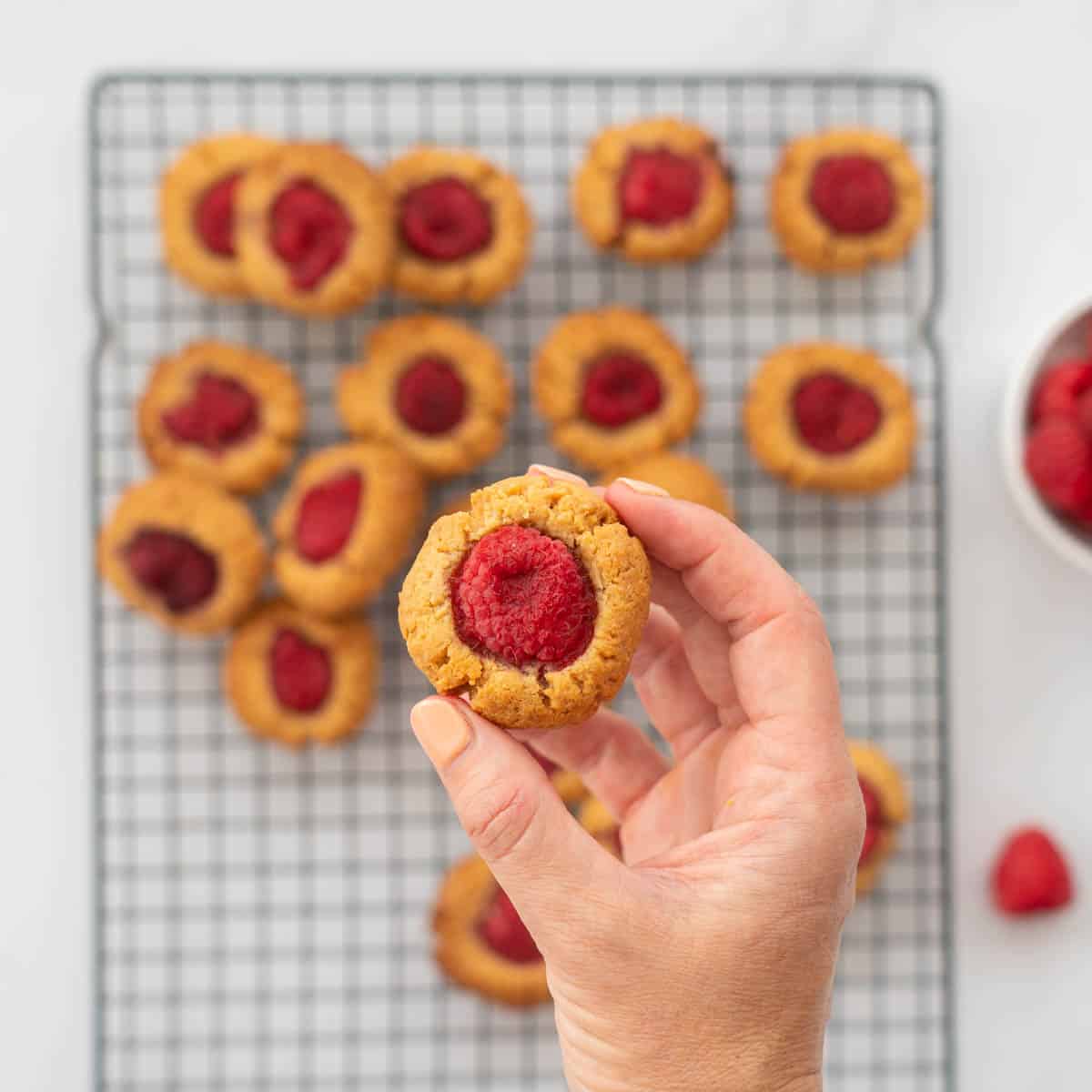 A thumbprint cookie topped with a raspberry being held above a cooling rack of other cookies.