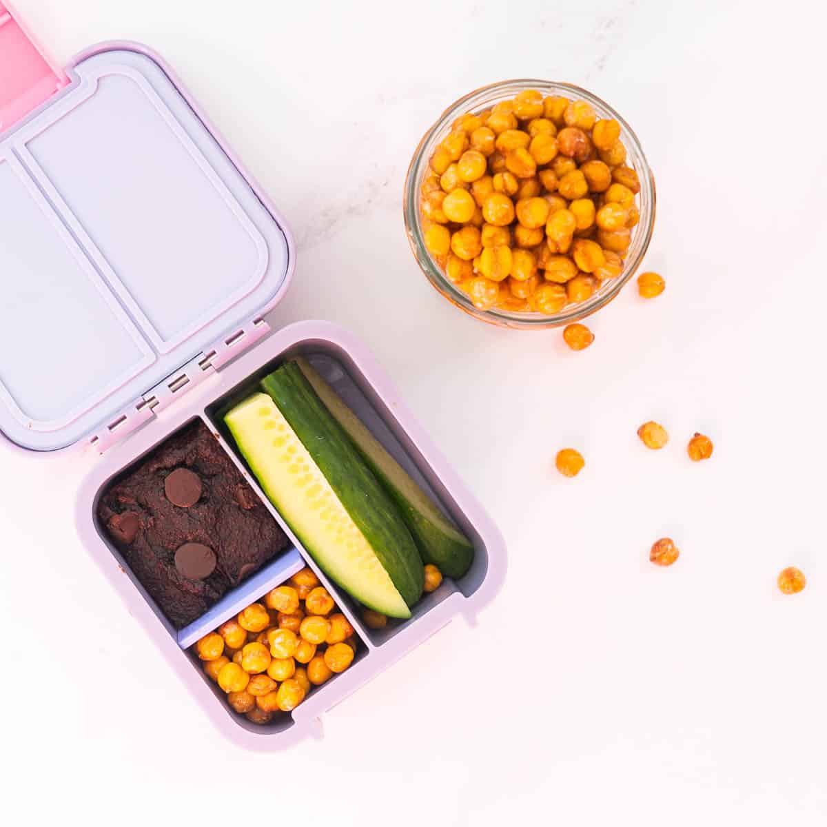 Roasted chickpeas in a lilac bento box with a piece of brownie and cucumber sticks.