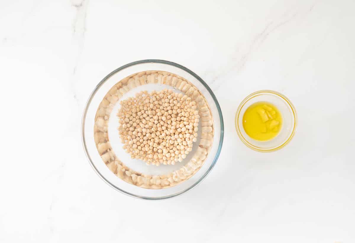 A large glass bowl of chickpeas soaking in water, next to a small bowl of garlic soaking in olive oil. 