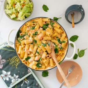 A large white casserole dish filled with salmon pasta bake on a table top with spinach leaves and a sliced lettuce salad.