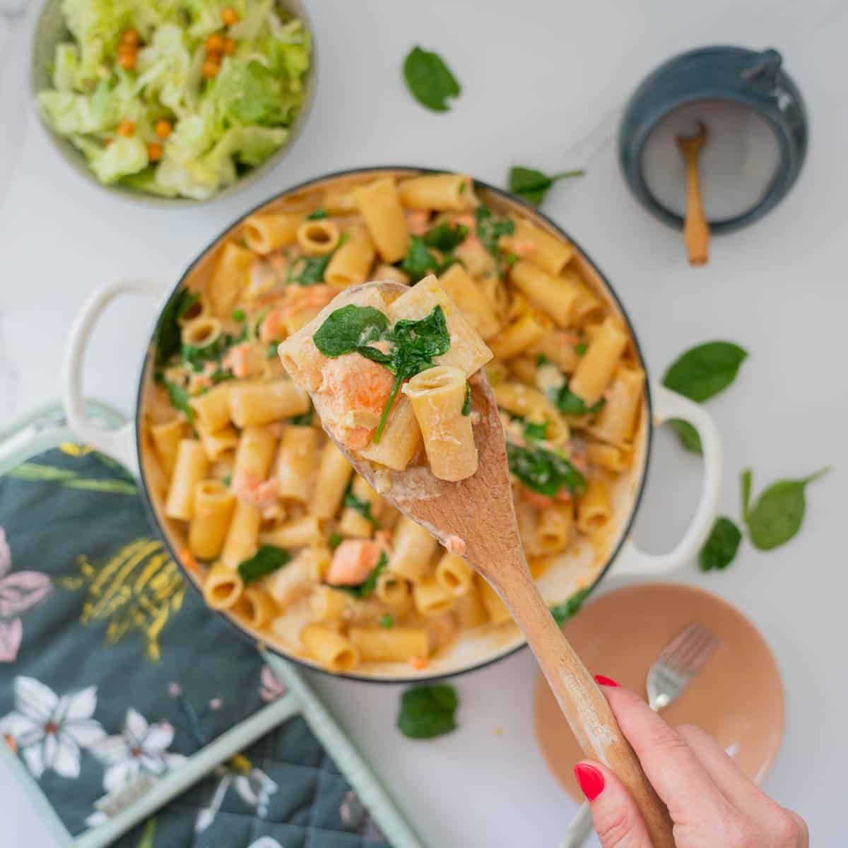 A spoonful of creamy  pasta, spinach leaves and cooked salmon being held above a large casserole dish of salmon pasta bake. 