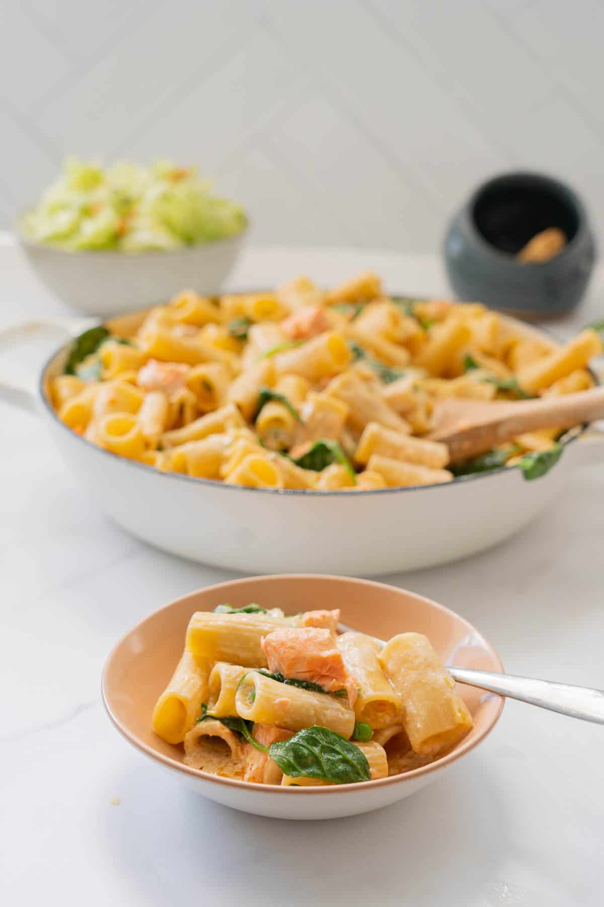 A light pink ceramic bowl  cooked pasta in a cremy sauce with salmon, spinach leaves and peas also visible.