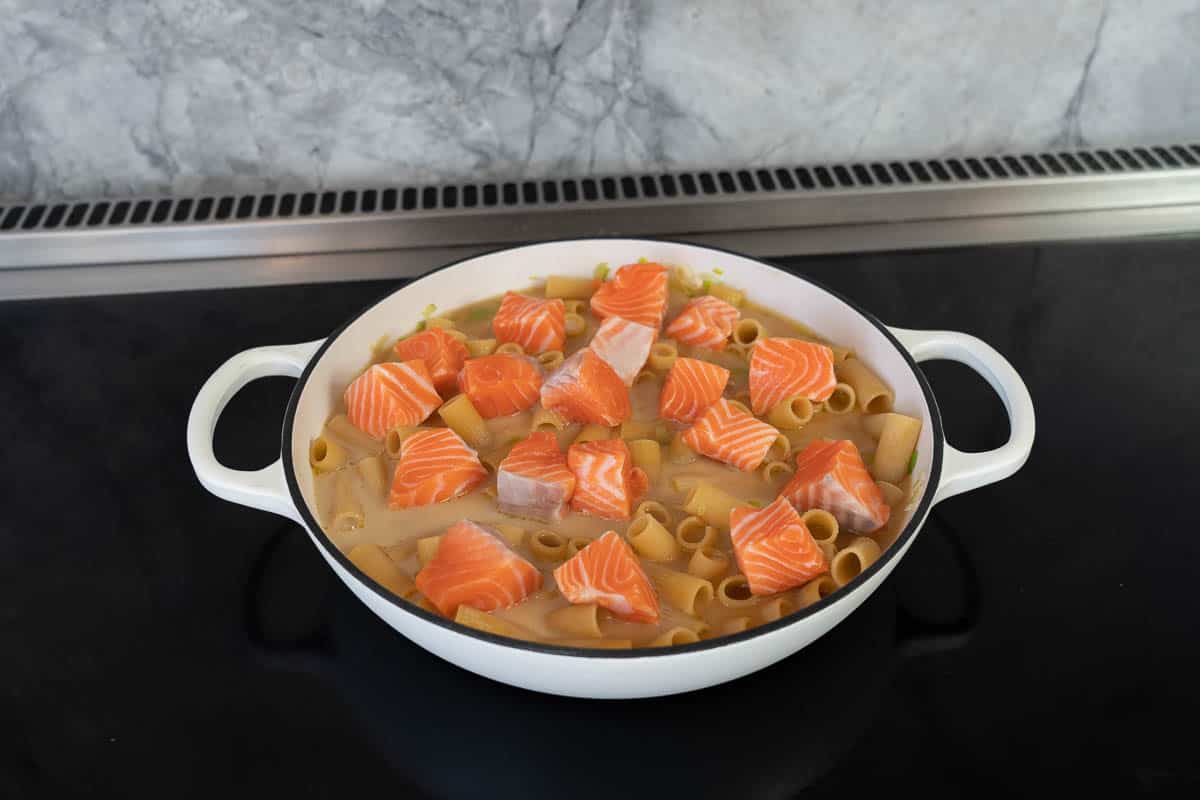 Cubes of salmon on top of pasta sitting in liquid in a large casserole dish on a stove top.