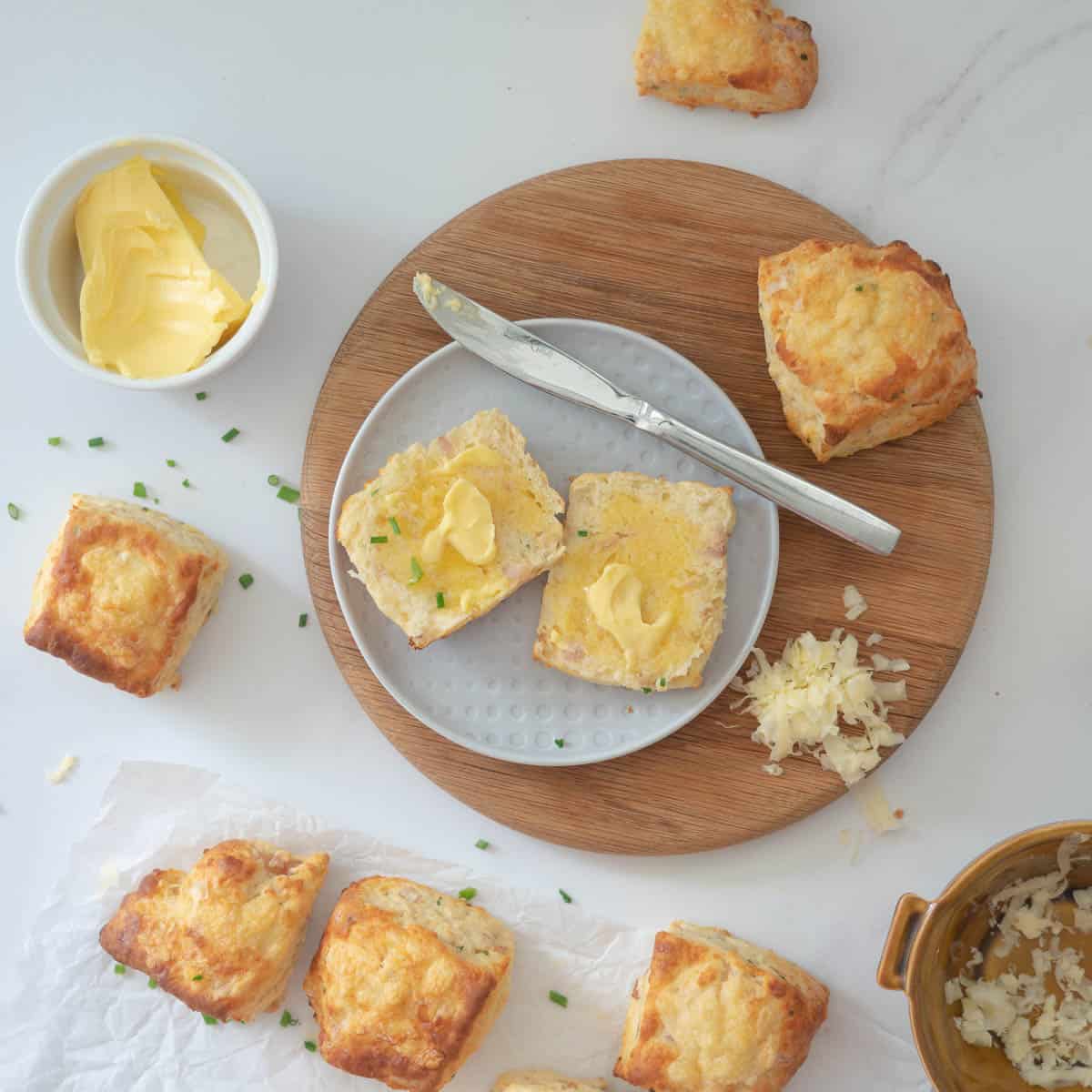 A scone split open and buttered on a wooden board surrounded by other scones and a small bowl of butter.