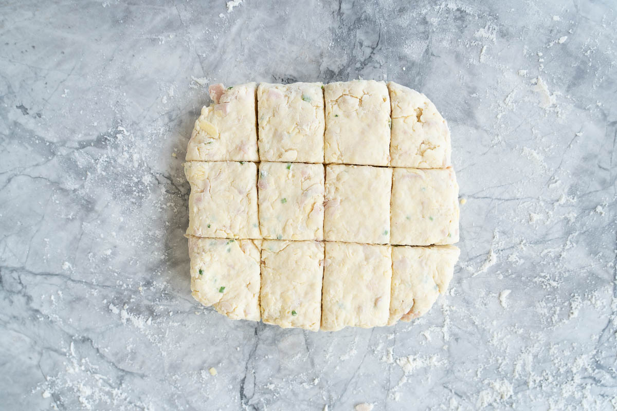 A square of scone dough, cut into 12 smaller squares on a grey marble bench top.