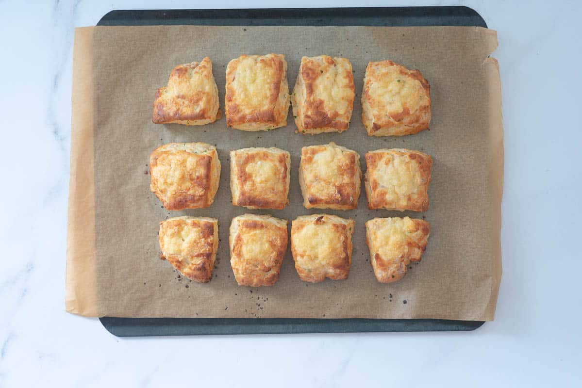 Twelve golden baked square scones topped with grated cheese on a lined baking tray.