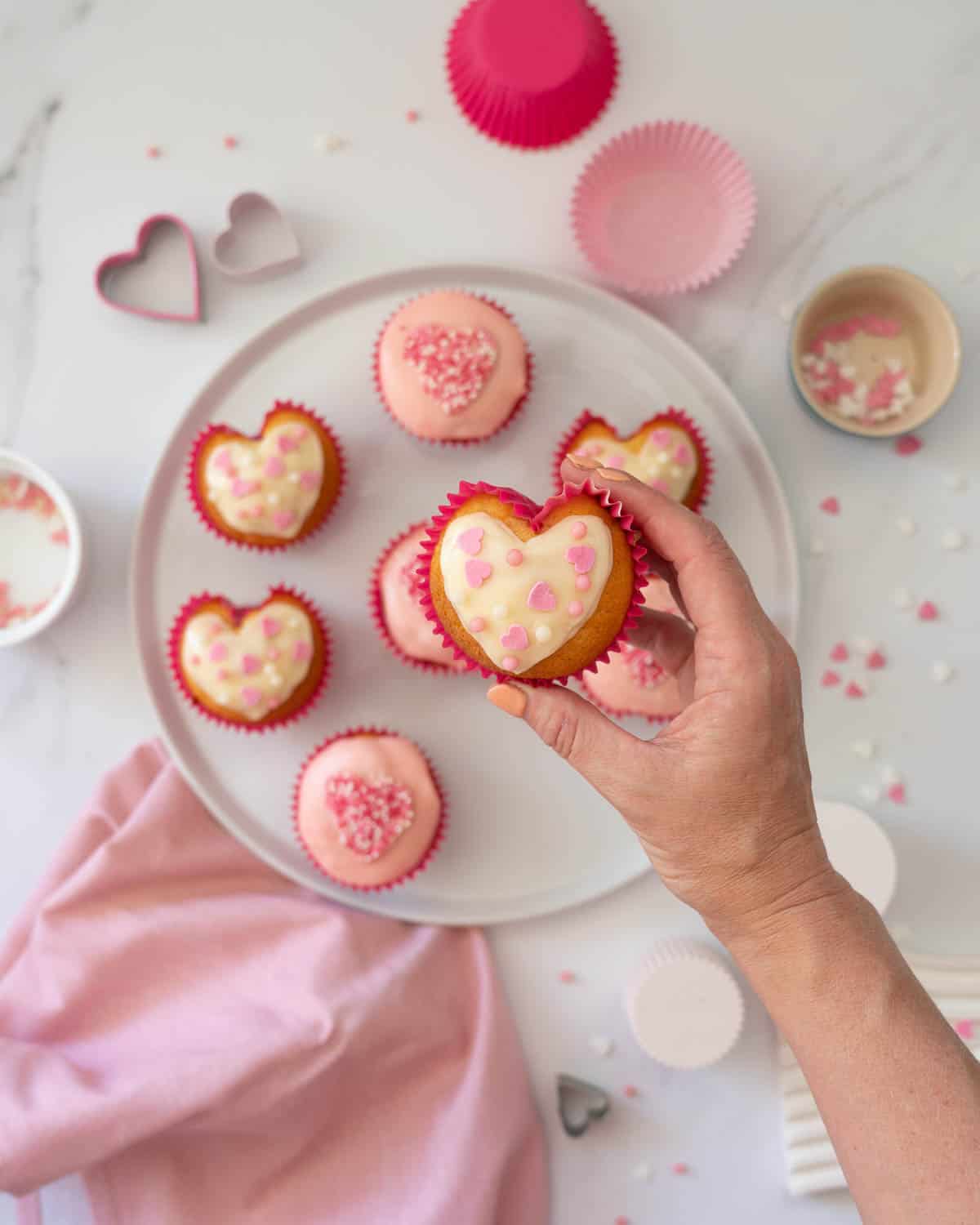 A heart shaped cupcake, frosted with white glaze, and topped with pink and white heart shaped sprinkles.