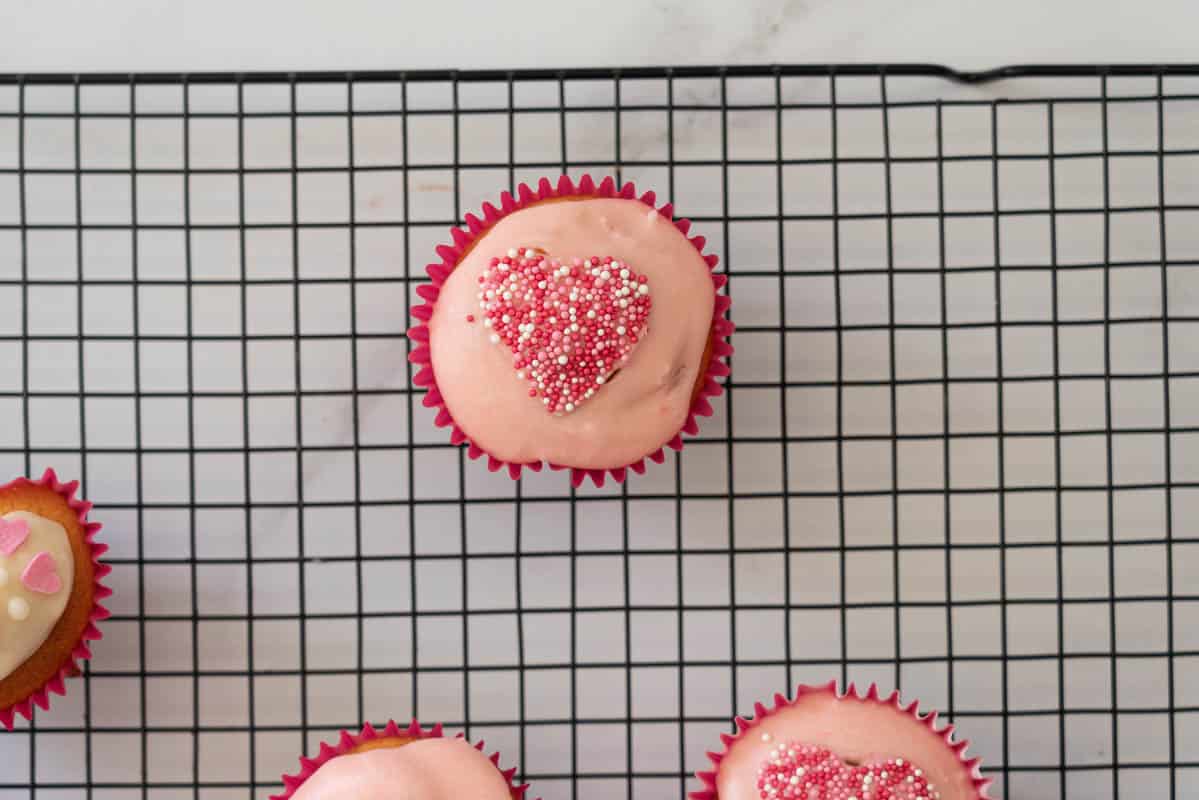 A cupcake glazed with pale pink glaze, and decorated with pink and white sprinkles in a heart shape. 