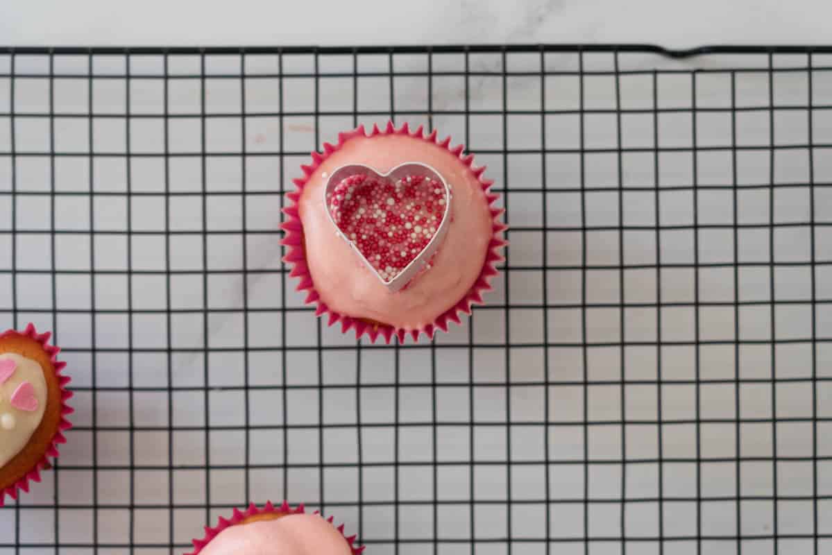 A glazed cupcake with a small heart cookie cutter sitting on top of it, the cutter is filled with sprinkles.