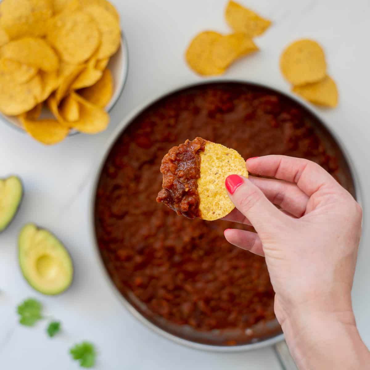 A corn chip loaded with bean based nacho mix, avocado and sour cream being held above a dinner table.