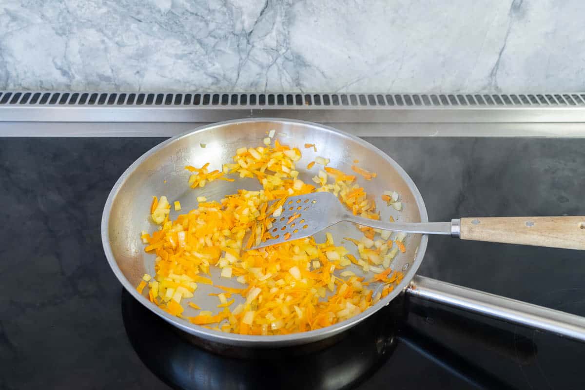 Sautéed diced onion and grated carrot in a stainless steel fry pan.