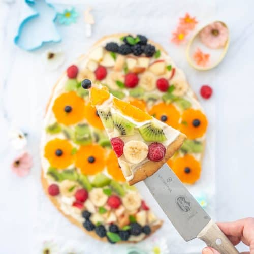 A slice of fruit pizza being held up above the entire fruit pizza on a bench scattered with Easter decorations