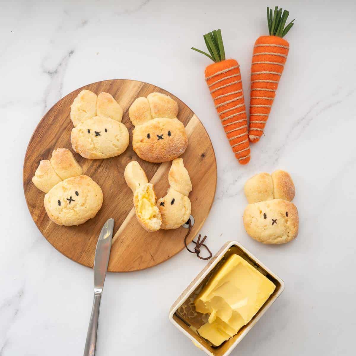 Four bunny shaped bread rolls on a round wooden board, one split in half and buttered.