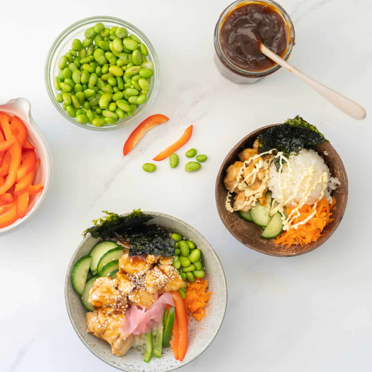 Two sushi bowls sitting on a table with a bowl of teriyaki sauce and edamame beans.