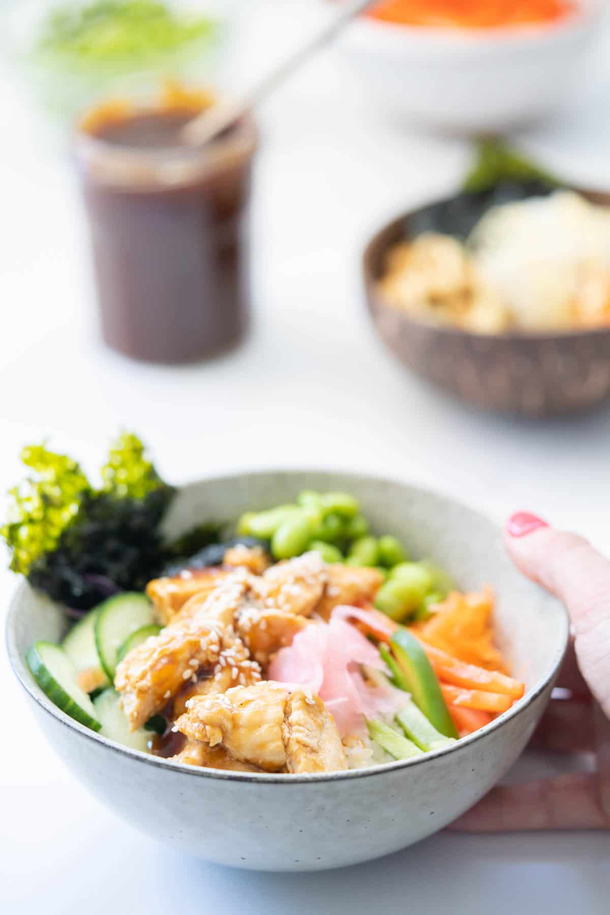 A bowl of teriyaki chicken Rice, vegetables and toasted seaweed pickled ginger and sesame seeds with a second bowl and jar of teriyaki sauce in the background.