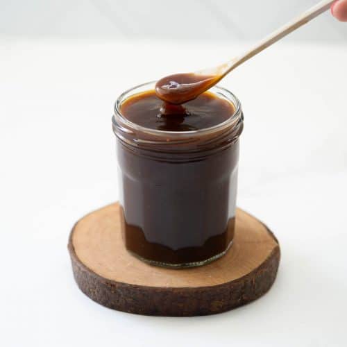 A spoonful of glossy brown teriyaki sauce coming being scooped out of glass jar.