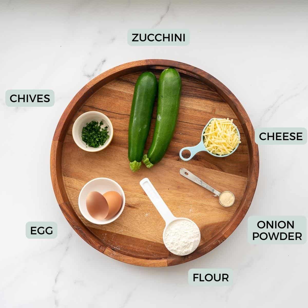 The ingredients to make zucchini tots on a round wooden tray with text overlay.