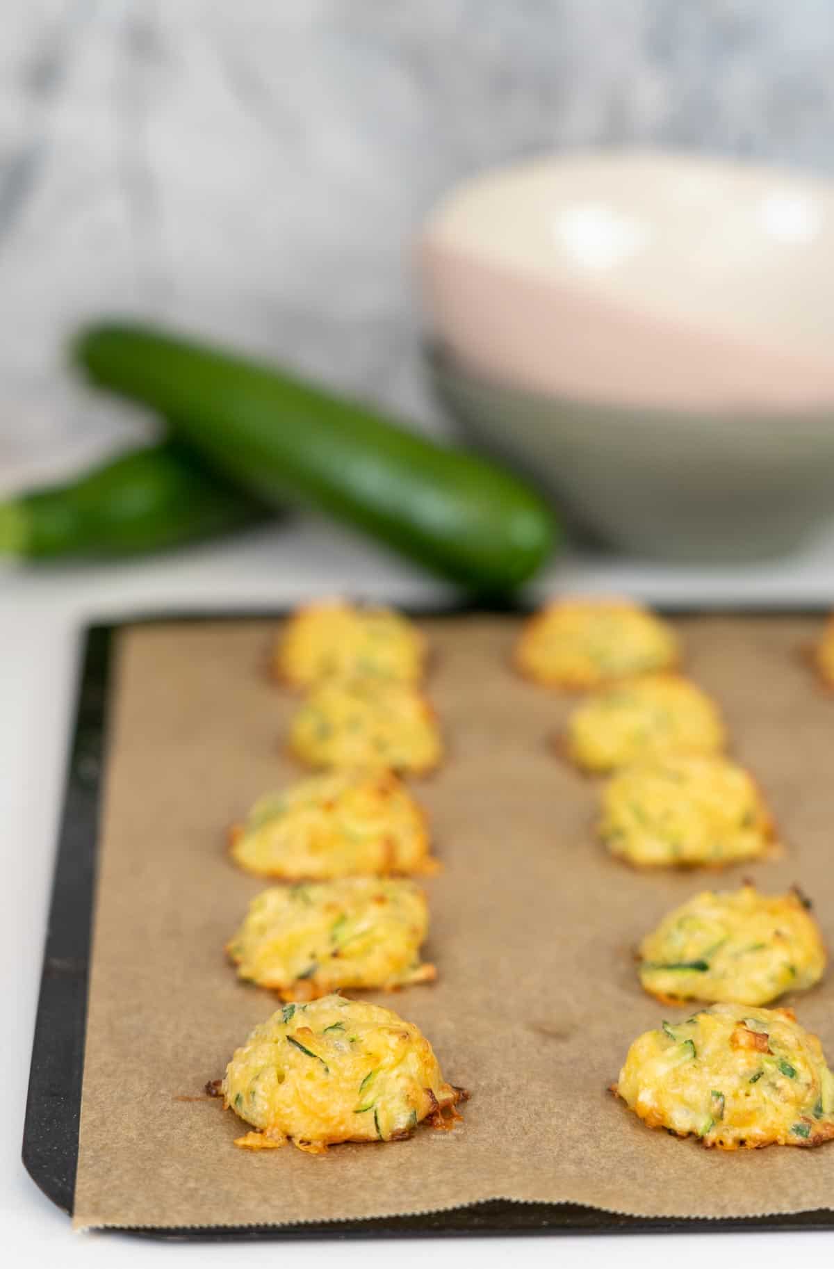 Zucchini tots on a lined baking tray with zucchini and coloured bowls in the background.