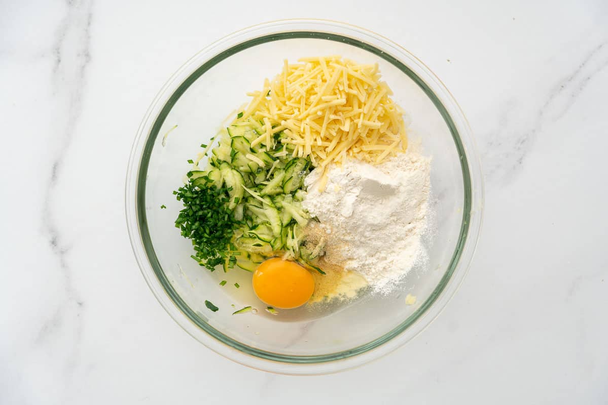 Grated zucchini, flour, grated cheese, chives, and egg in a glass mixing bowl.