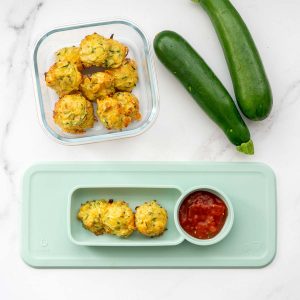 Three zucchini tots on a silicone feeding mat with a red dipping sauce, next to a glass container filled with zucchini tots.