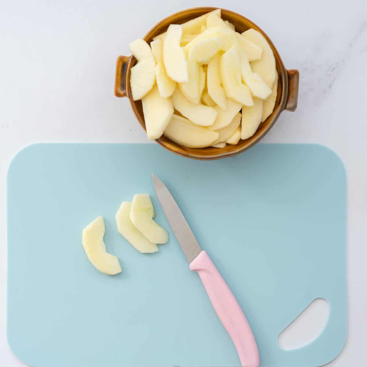 Peeled and cored apple slices in a brown bowl, next to a light blue chopping board and pink handled paring knife.
