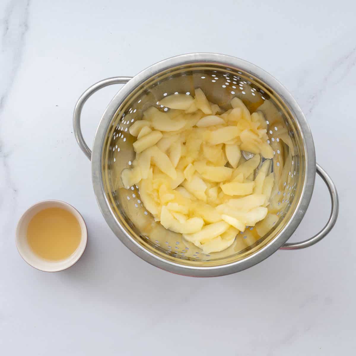 Cooked apple slices in a colander next to a small bowl of apple water.