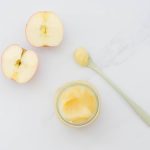 Apple puree in a small glass jar, with a spoonful of puree and halved apples on the bench next to the jar.