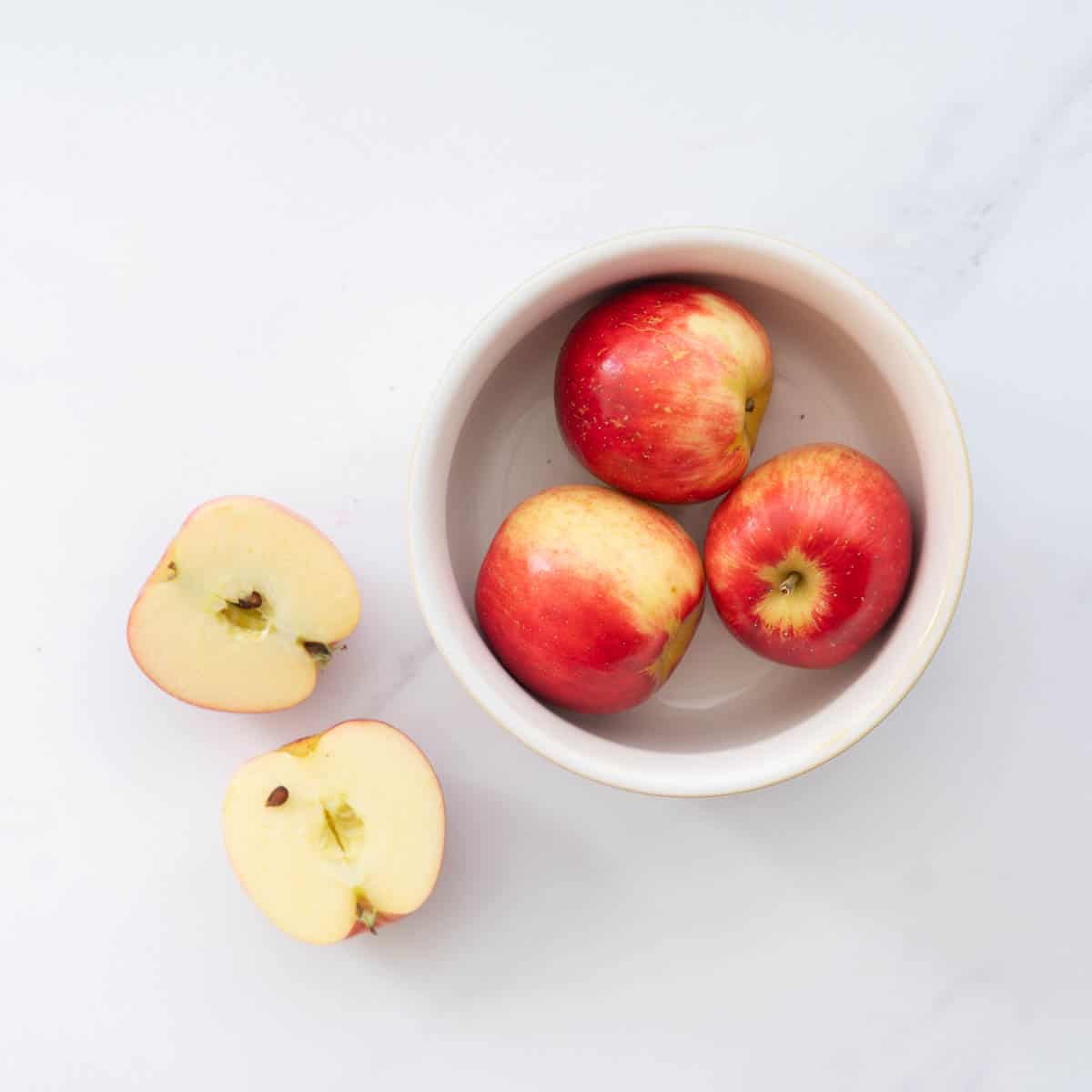 Three red skinned apples in a small bowl, next to a haved apple on a light marble bench.
