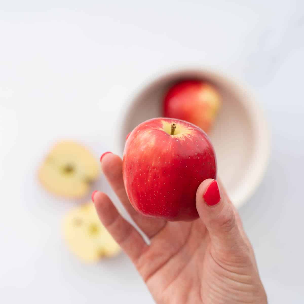 A ripe red skinned apple being held above a bowl of apples.