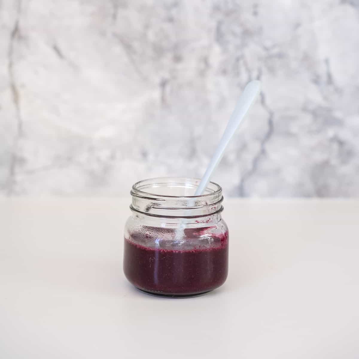 Purple blueberry puree in a small glass jar with a blue bamboo spoon in front of a marble splash back.