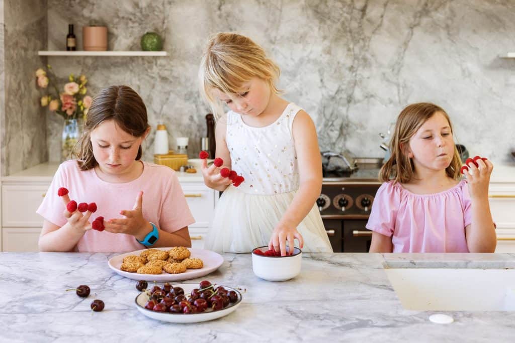 Three girls in a kitchen eating raspberries of their hands