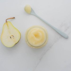 A glass jar of pear puree next to half a pear and a pre-loaded spoon of pear puree on a bench.