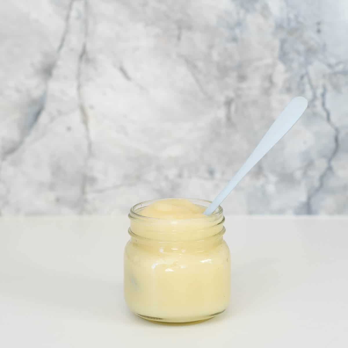A glass jar of pear puree with a blue bamboo spoon sitting in-front of a marble splashback.