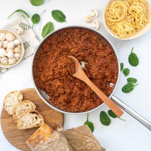 A large fry pan of bolognese sauce on a table set with cooked pasta, fresh bread and spinach salad.