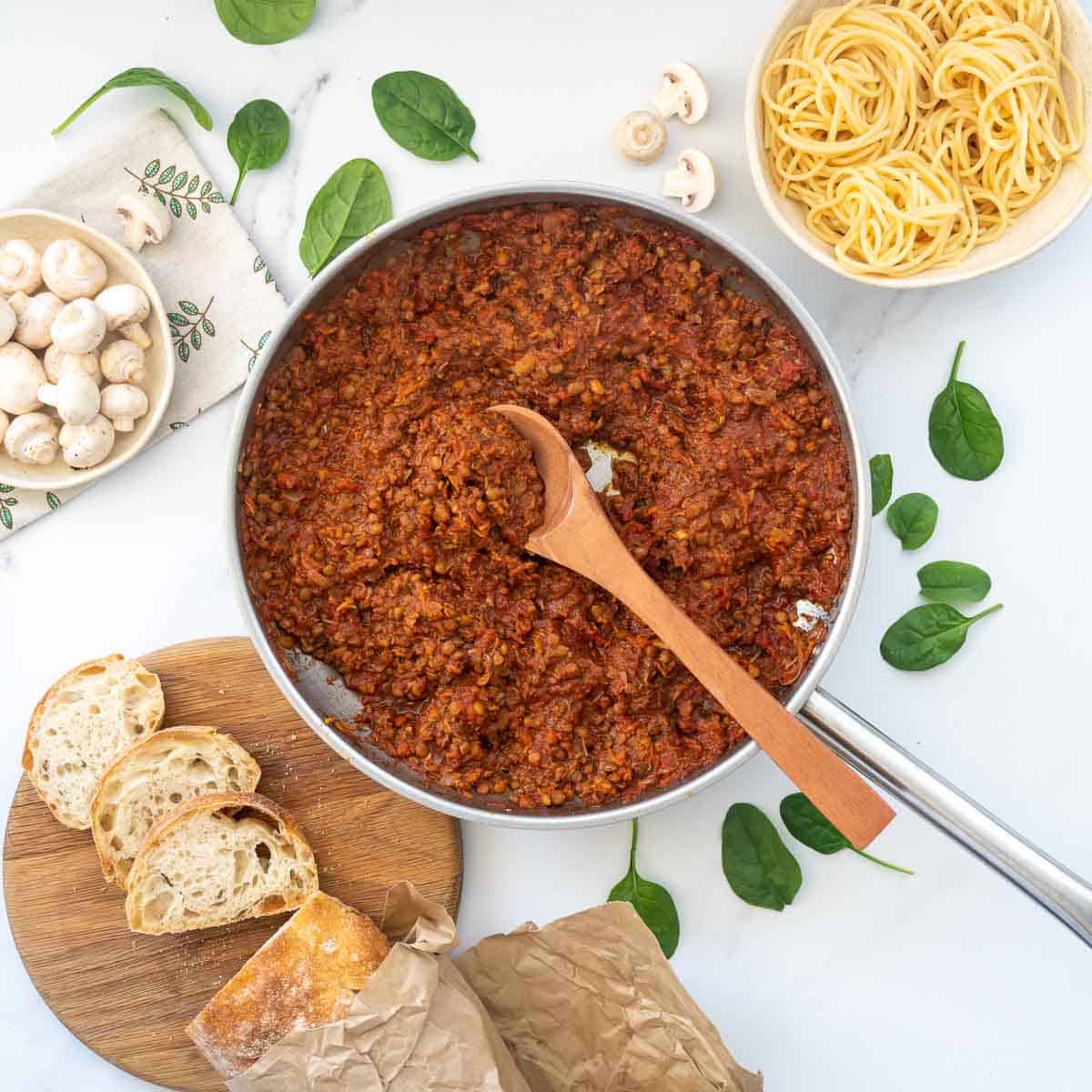 A lentil based pasta sauce in a large fry pan with bread and cooked spaghetti. 