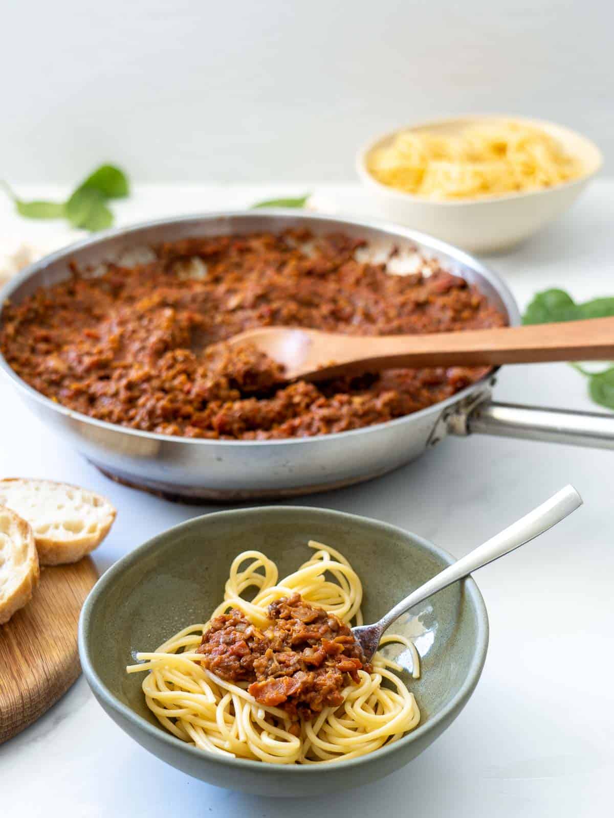 A green ceramic bowl of spaghetti bolognese on a table in front of a large fry pan of bolognese sauce. 