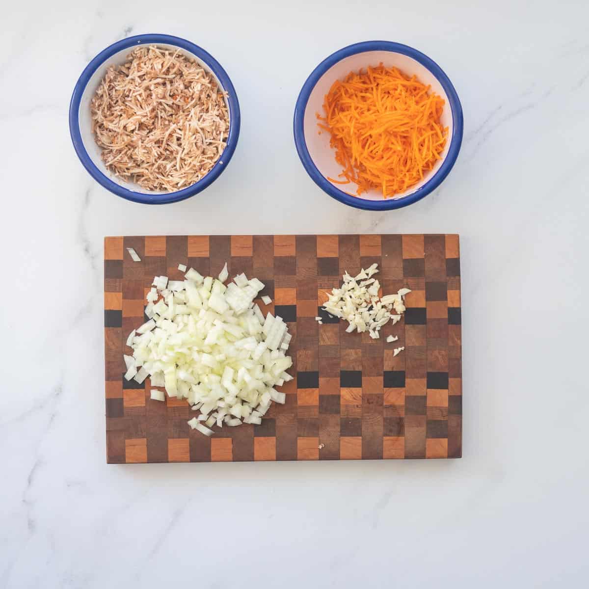 Bowl of grated vegetables and a chopping board with diced onion and crushed garlic cloves.