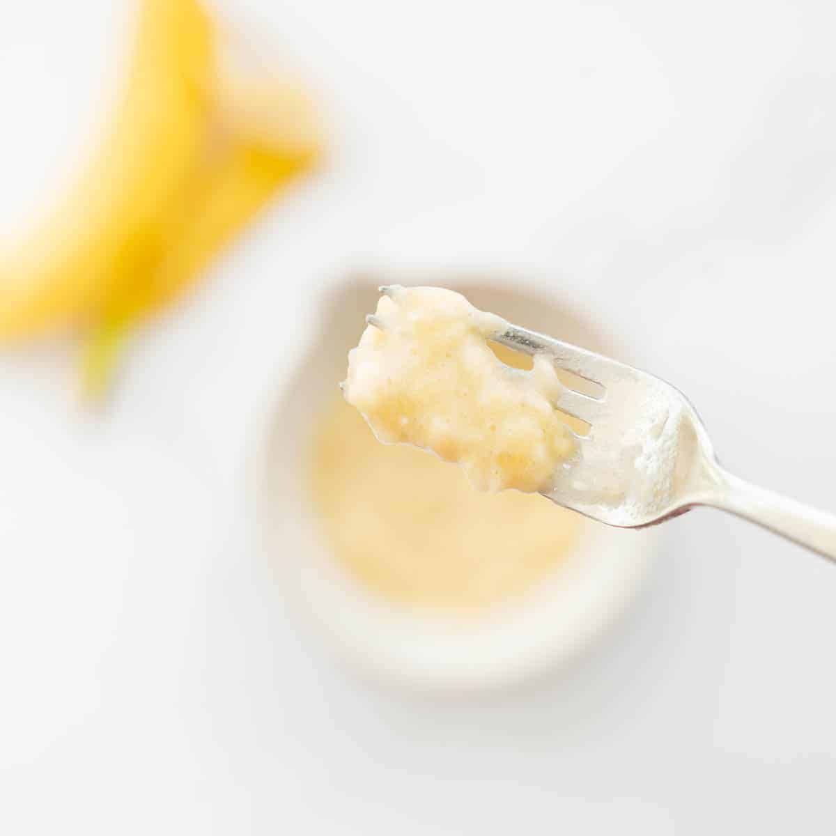 In the faded background you can see a banana skin and a bowl. Up close is a fork with creamy mashed banana