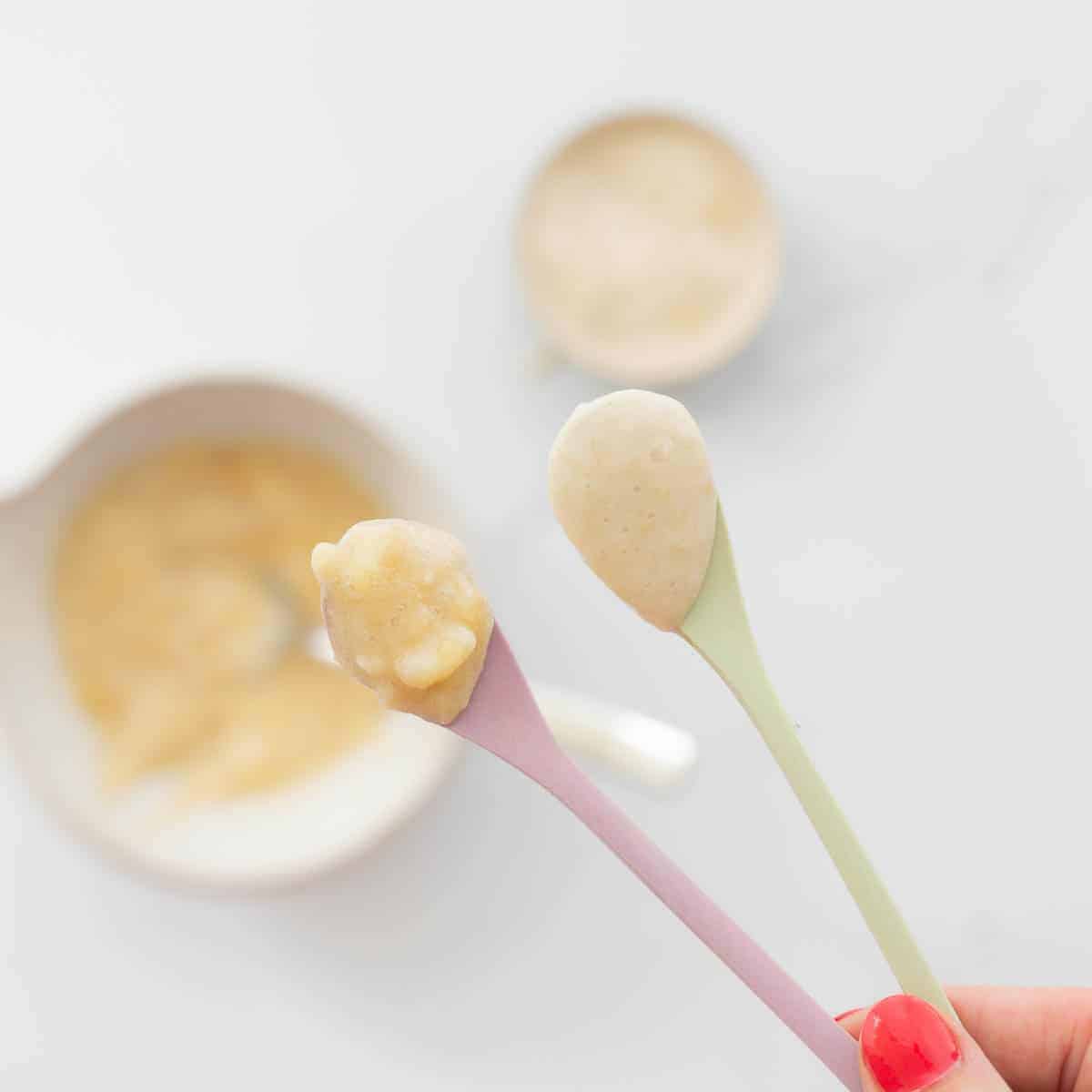 Two baby spoons being held up to the camera, one of mashed banana, and one of banana puree. 