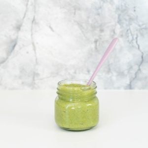 A glass jar of green puree sitting in front of a grey marble splashback.