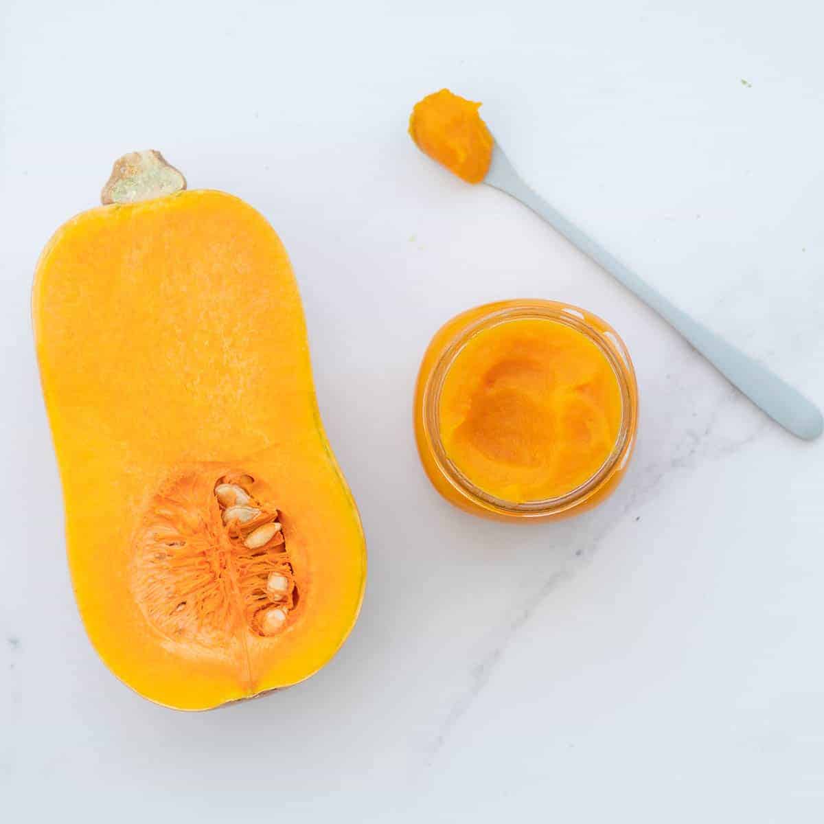 A butternut cut in half beside a jar of smooth orange puree with a blue spoon next to the jar with puree on it