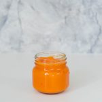 A glass jar of orange puree sitting in front of a grey marble splashback.